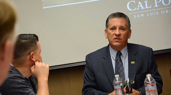 Man in suite in front of a screen who is talking. There are two other people in the photo who are paying attention to him speak.