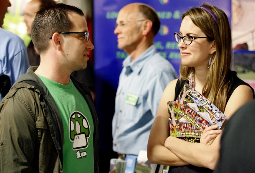 Two people, a man and a woman, both wearing glasses, talking with each other in a group setting with other people.