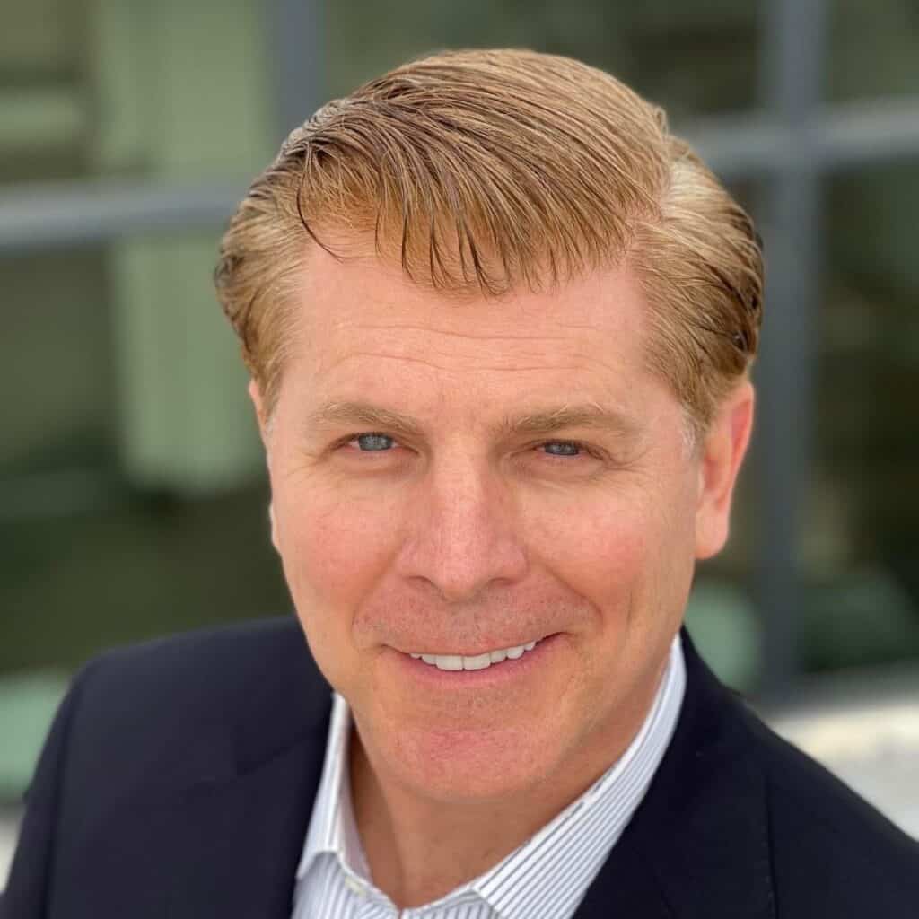 Patrick smiles at the camera, which is cropped to show only his face and shoulders. He has red hair and is wearing a dark blue suit jacket with striped shirt.