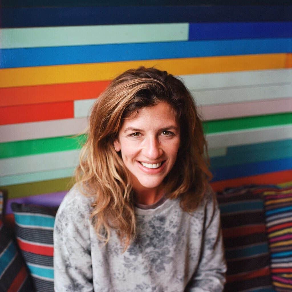 Leah is smiling at the camera in front of a colorful stripy wall. She is wearing a gray sweatshirt and has blonde hair.