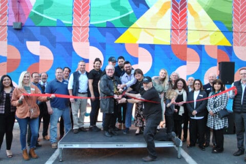 A large group of people stand in front of a colorful mural, smiling at the camera. The person in the middle is holding oversized scissors that say "San Luis Obispo Chamber of Commerce" and is about to cut a red ribbon stretched across the group of people.