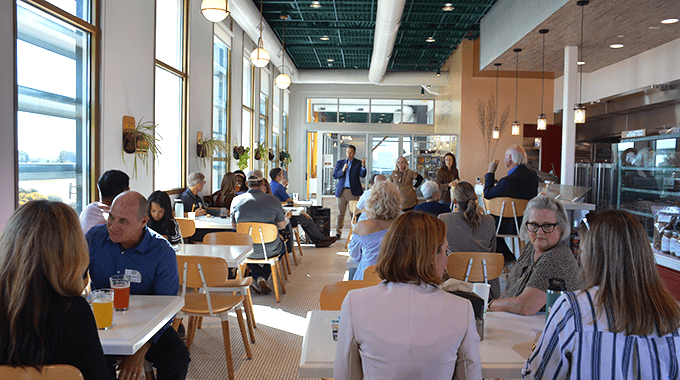 Group of people sitting at small tables and chairs in groups of two and four in a restaurant like setting. They are listing to a male speaker, Jim Dantona, who has a microphone and is talking. Setting is bright and modern.