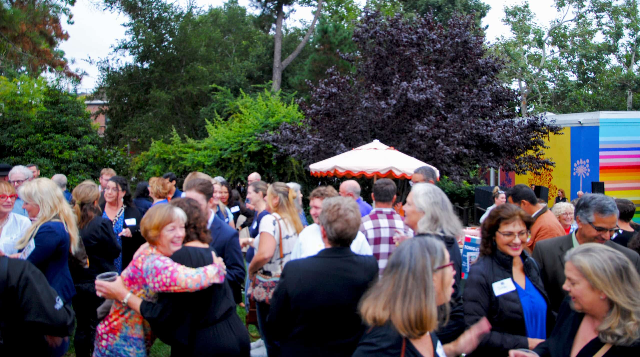 Set in the outdoors on the lawn area of the SLO Museum, mix of people gathered and mingling, talking, hugging and enjoying the space. The background are trees and off to the right is a colorful corner of the SLO Museum painted in bright colors.