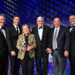 Six people on a blue silver stage dressed formal. There is a woman in the middle wearing a gold animal print jacket holding a round glass award. The men are all wearing tuxedo's or suits. They are all smiling towards the camera.