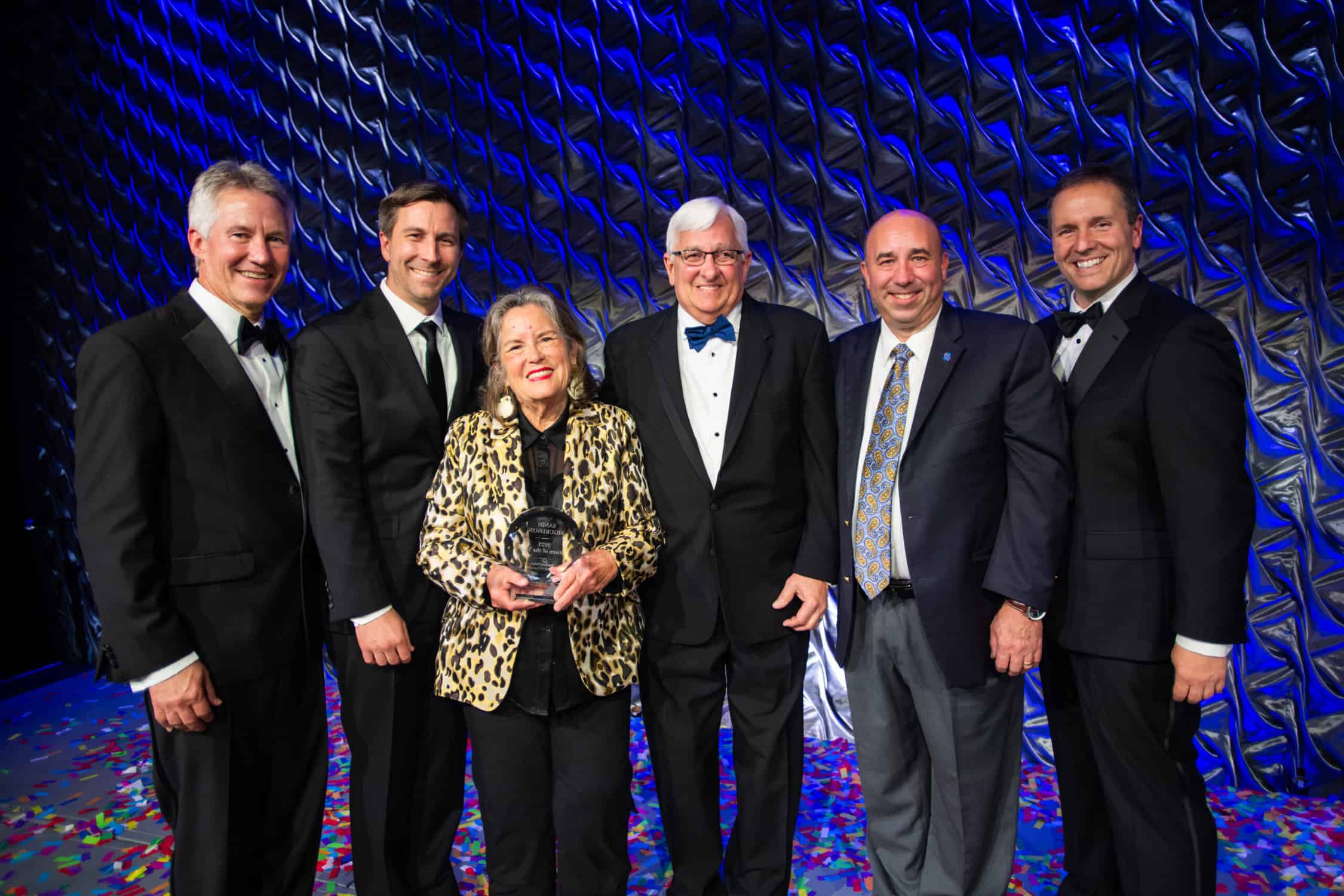 Six people on a blue silver stage dressed formal. There is a woman in the middle wearing a gold animal print jacket holding a round glass award. The men are all wearing tuxedo's or suits. They are all smiling towards the camera.