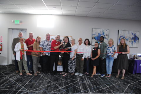 Group of people holding a red ribbon with one person in the middle holding oversized plastic scissor to mock cut ribbon. All are smiling toward the camera, set inside a conference room with dark carpeting.