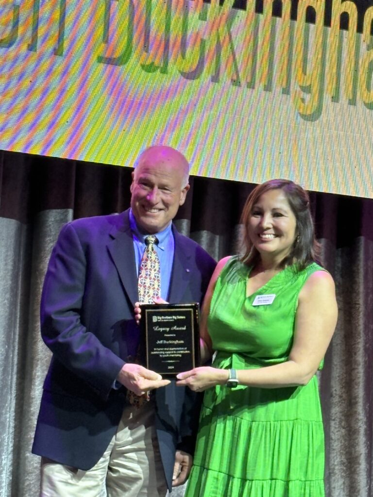 Man in blue suit, Jeff Buckingham from Astound Business Solutions, is standing next to and receiving 2024 "Legacy Award," from woman in green dress, Stacy Salame, of Big Brothers Big Sisters for SLO County.