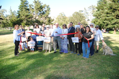 Large group of people, kids and adults, gathered in sunny park setting for group photo with political candidate.