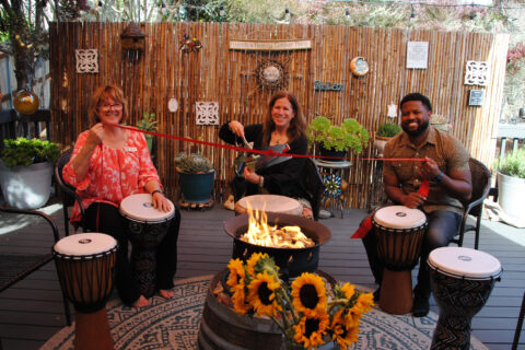 Three people are sitting with large drums in front of them along with a fire like a campfire, The middle woman is holding large oversized fake scissors pretending to cut a red ribbon that isheld by the other twp people on either side of her; one a woman and the other a man.
