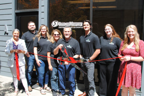 Group of people in front of a business called Sousa and Company, CPS's LLP. They are all wearing black company shirts and the person in the middle is holding oversized scissors for a ribbon cutting. They are all smiling for the camera.