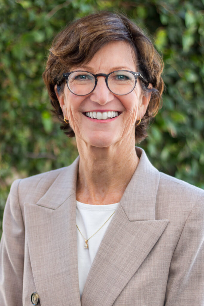 A picture of a woman with short brown hair wearing classes in a beige shirt and tan jacket smiling at the camera.