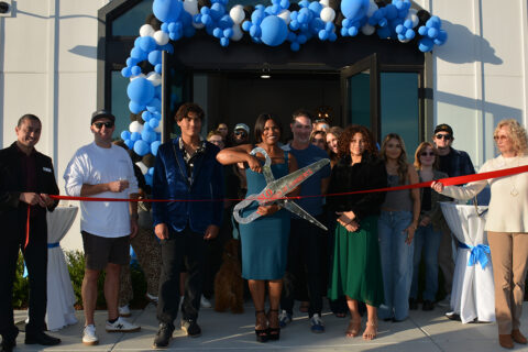 Group of people taking a posed photo for a ribbon cutting ceremony with main person holding fake large scissors getting ready to cut red ribbon.